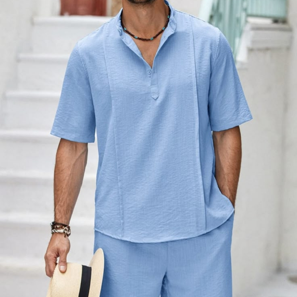 A man wearing a light blue, short-sleeved, collared shirt and matching shorts, holding a straw hat with a black band, standing outdoors near a white staircase and railing.
