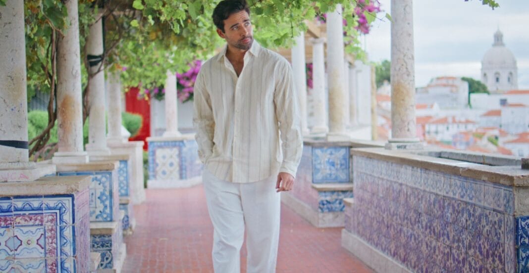 A man dressed in light-colored, relaxed clothing walking along a decorative outdoor colonnade with colorful tiles and greenery, overlooking a scenic view of a town with domed buildings.