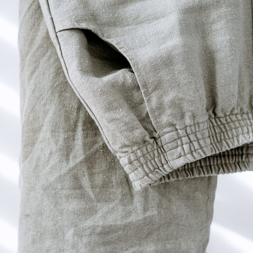 A close-up of a beige fabric garment with a visible pocket and elastic ruffle at the hem, showing textured, slightly wrinkled material.