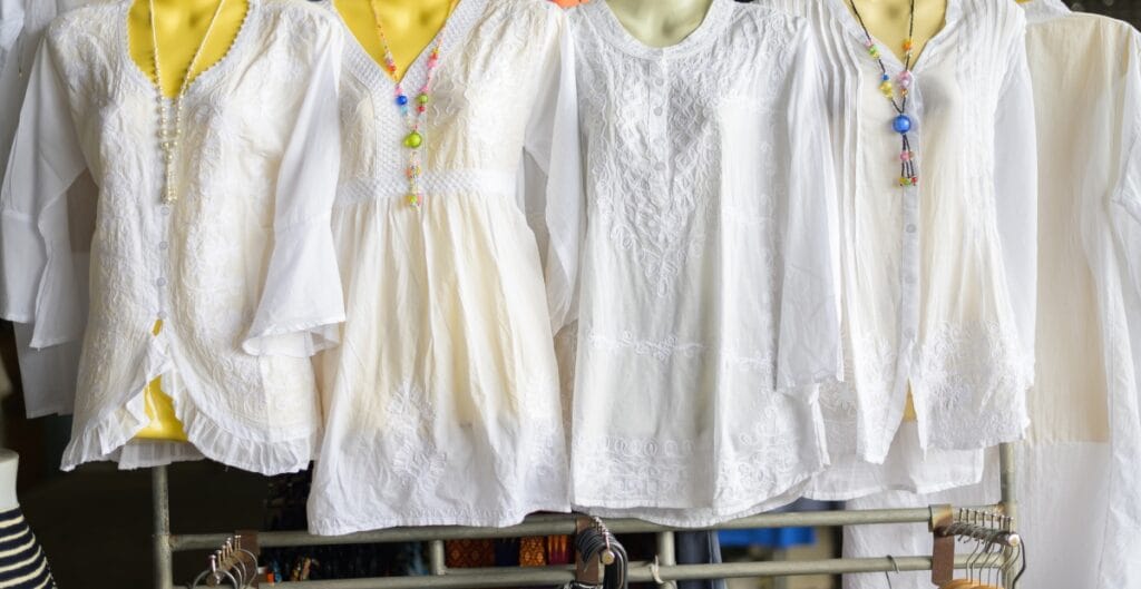 A row of white embroidered blouses hanging on a clothing rack, with some featuring colorful necklaces, set against a yellow background.