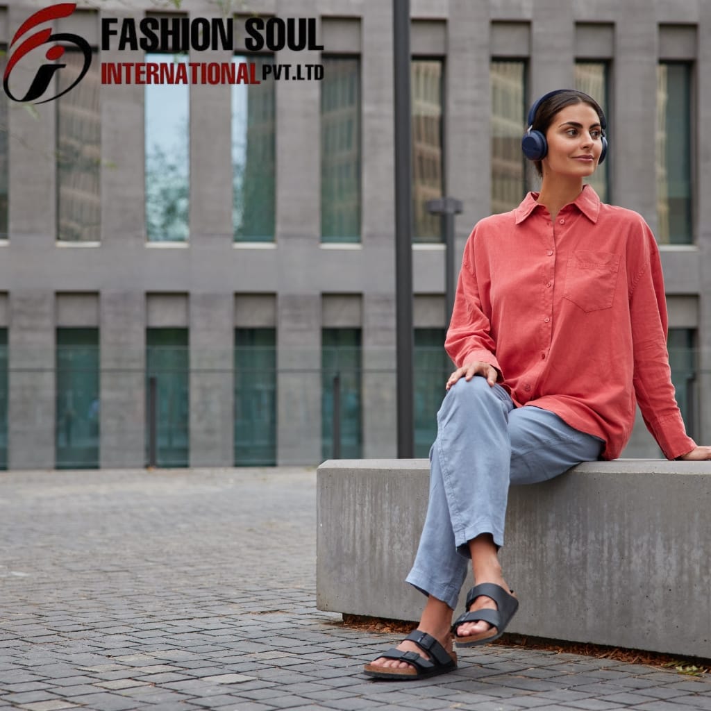 A woman sitting on a concrete bench in an urban setting, wearing a coral-colored shirt, light blue pants, black sandals, and large headphones, with a modern building in the background.
