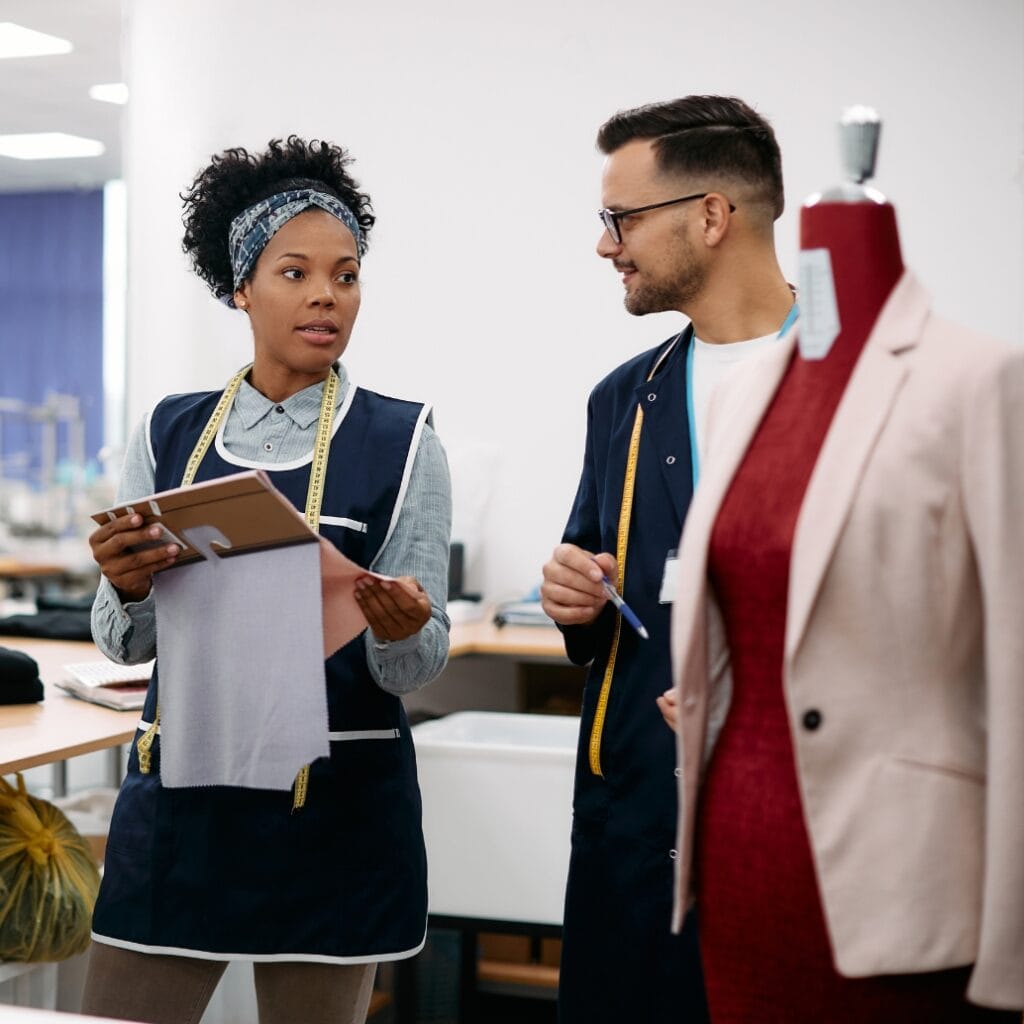 Three individuals are engaged in a conversation in a professional setting, with one woman holding a fabric sample and a clipboard, and two men listening and taking notes. A mannequin wearing a beige blazer and red top is visible in the foreground on the right.