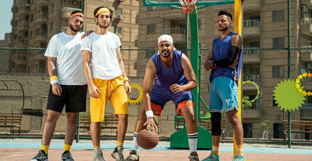 Group of five men wearing colorful sportswear posing on an outdoor basketball court, with a basketball and gym equipment in the background.