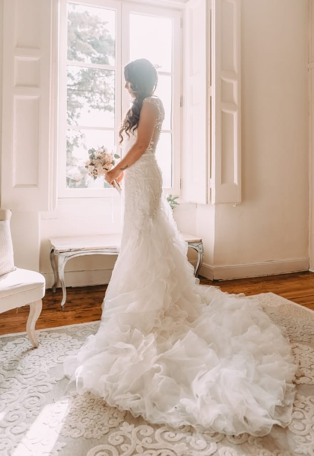 A bride standing indoors by a large window, wearing a stunning white mermaid-style wedding gown with ruffled, layered fabric cascading into a dramatic train.