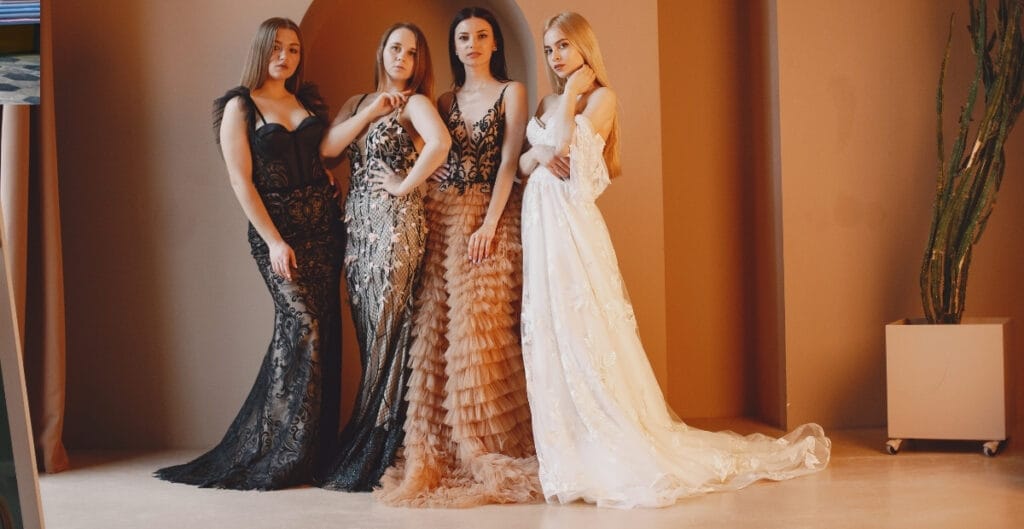 Four women standing side by side, each wearing elegant mermaid-style gowns in different designs and colors, in a studio with a warm background and a potted plant on the right.