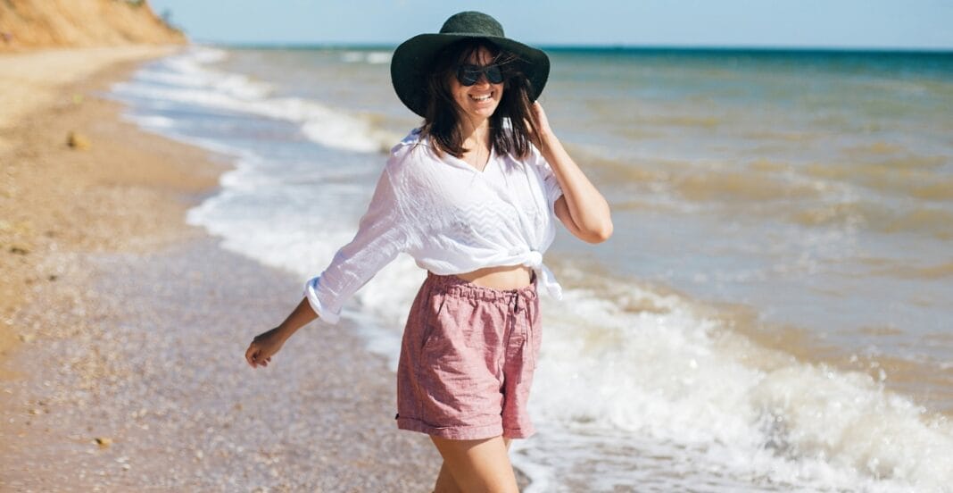 A woman wearing a large black sunhat, sunglasses, a white knotted blouse, and pink shorts, smiling and enjoying a walk along the beach with gentle waves behind her.