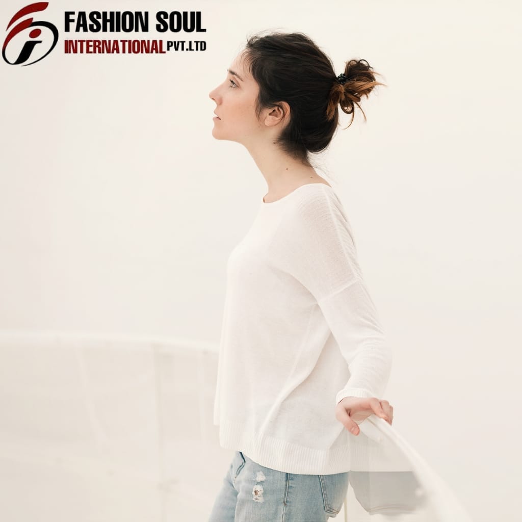 A young woman with dark hair tied in a messy bun, wearing a white sweater and ripped jeans, stands in profile against a plain, light-colored background. She holds a transparent object with her right hand, which appears to be a curtain or a similar fabric.