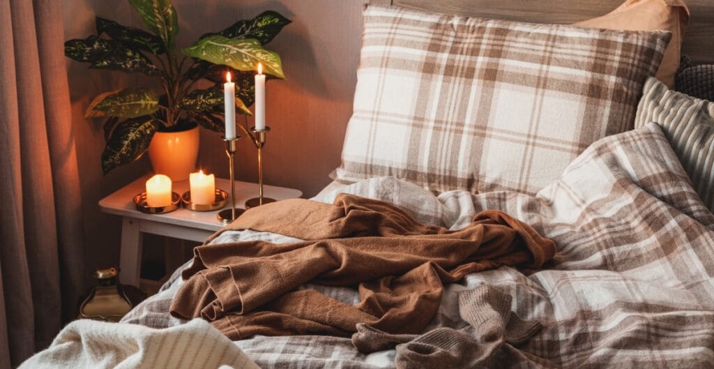 A cozy bed with plaid bedding, partially unmade, next to a small white nightstand with lit candles and a potted plant in a warm-lit room.