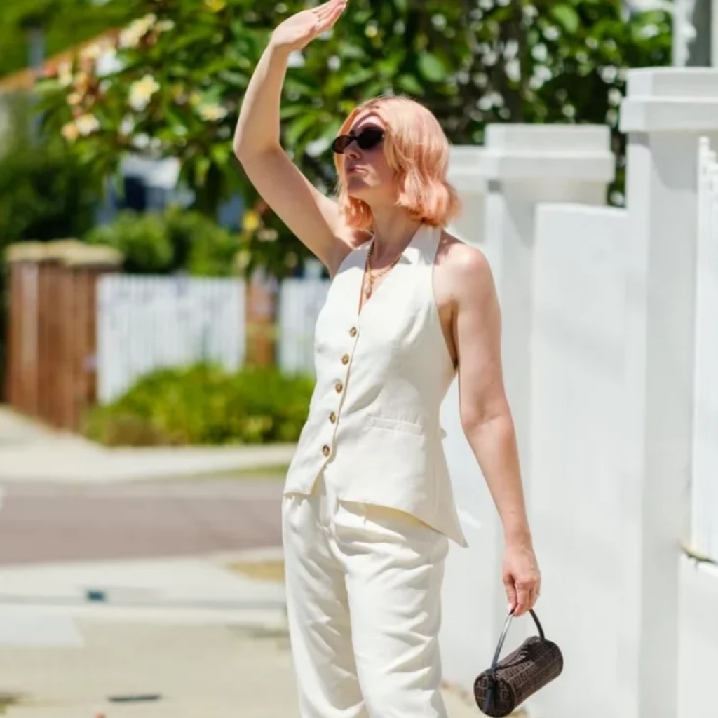 A woman with light hair and wearing sunglasses, dressed in a sleeveless cream-colored top and matching pants, standing outdoors on a sunny day. She is holding a small cylindrical bag in one hand and shielding her eyes with her other hand, as she looks into the distance. Green foliage and a white fence are visible in the background.