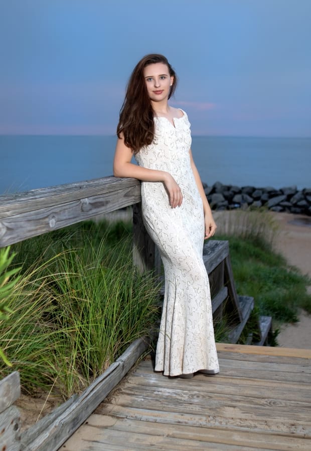 A young woman in a white, elegant mermaid gown standing on a wooden boardwalk by the beach at sunset. She has long, wavy brown hair and a subtle smile, leaning casually against the railing with her hand resting on it. The ocean and a rocky shoreline are visible in the background.