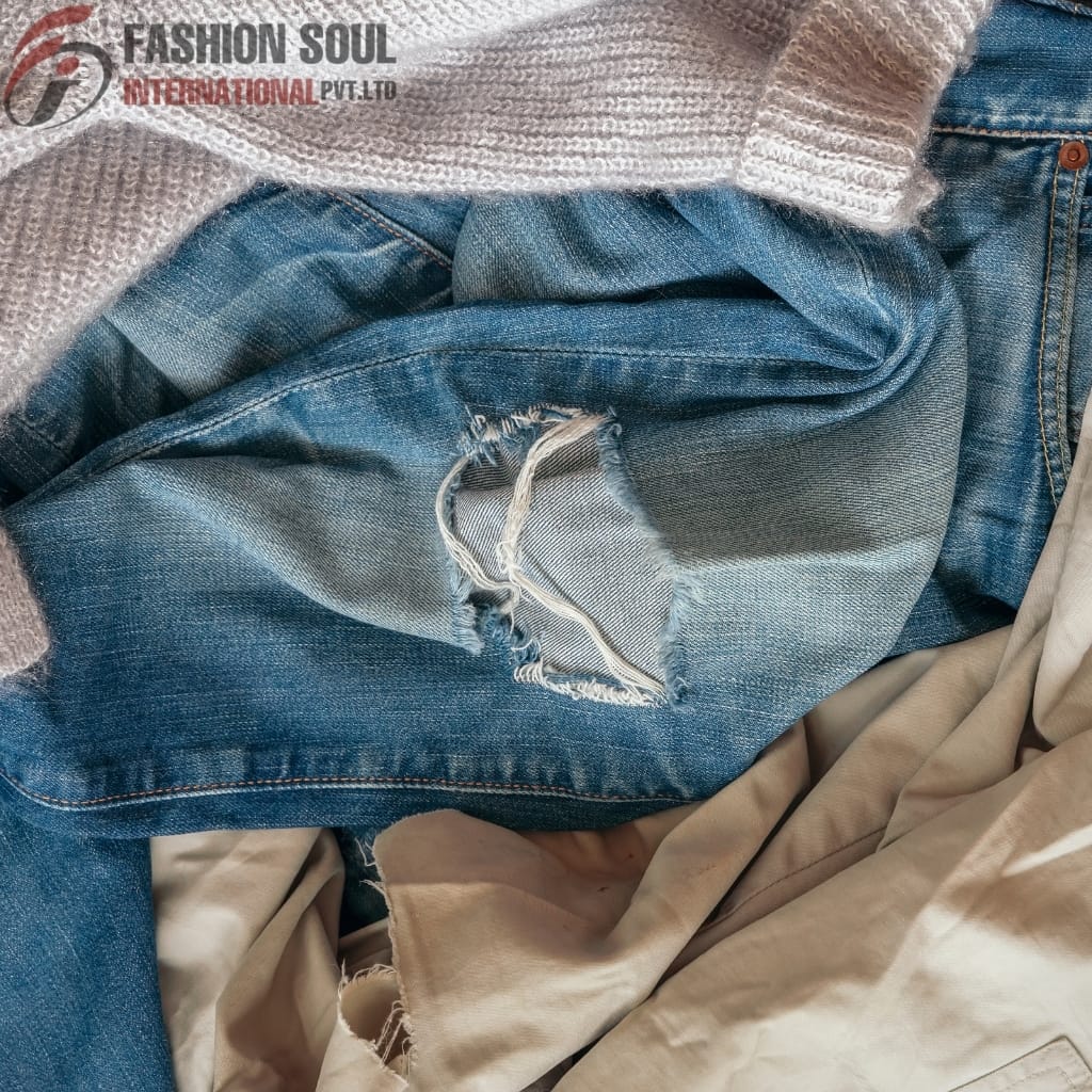 Close-up of layered clothing with distressed denim jeans, a beige fabric, and a textured gray sweater, with the "Fashion Soul International" logo in the top left corner.