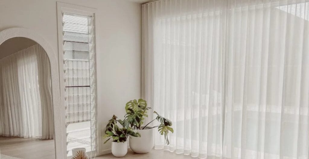 Bright interior corner with sheer white curtains, a large window, and three potted houseplants near the window.