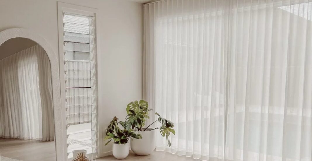 Bright interior corner with sheer white curtains, a large window, and three potted houseplants near the window.