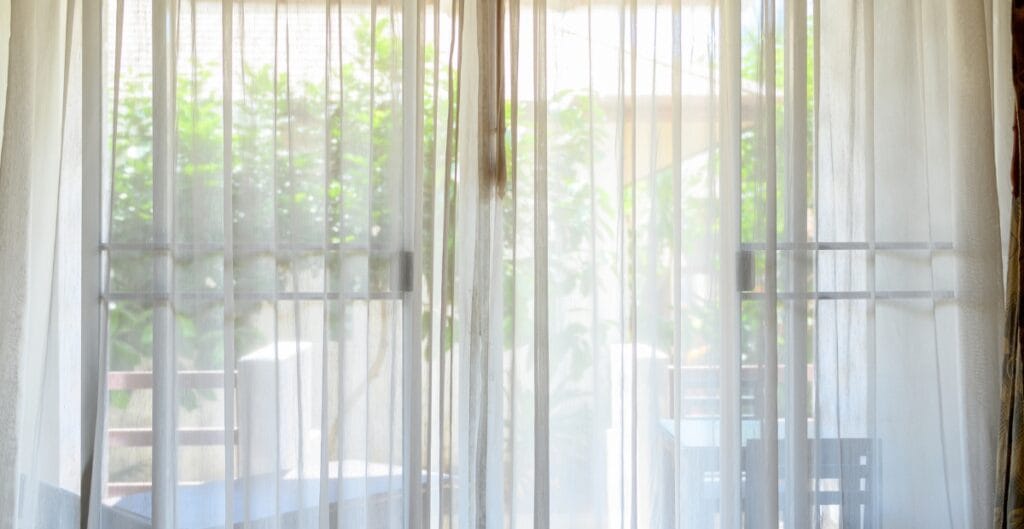 A view through sheer white curtains gazing out onto a patio with a table and chairs surrounded by greenery.