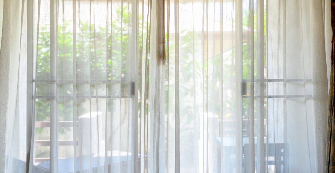 A view through sheer white curtains gazing out onto a patio with a table and chairs surrounded by greenery.