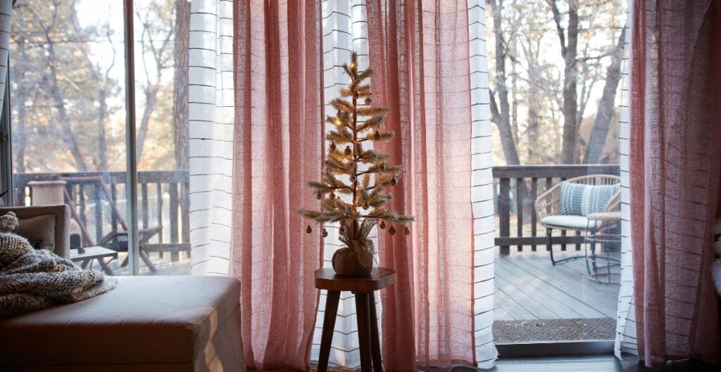 A cozy interior scene featuring a small decorated Christmas tree on a stand, placed in front of pink and white curtains that open to a wooden deck with outdoor furniture and trees in the background.