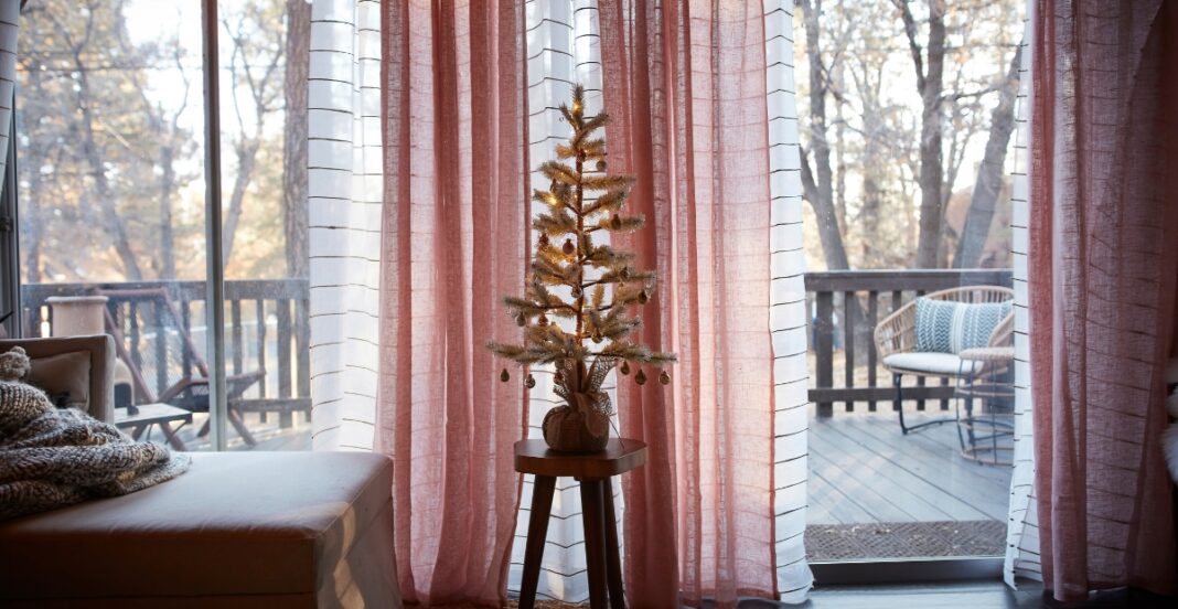 A cozy interior scene featuring a small decorated Christmas tree on a stand, placed in front of pink and white curtains that open to a wooden deck with outdoor furniture and trees in the background.