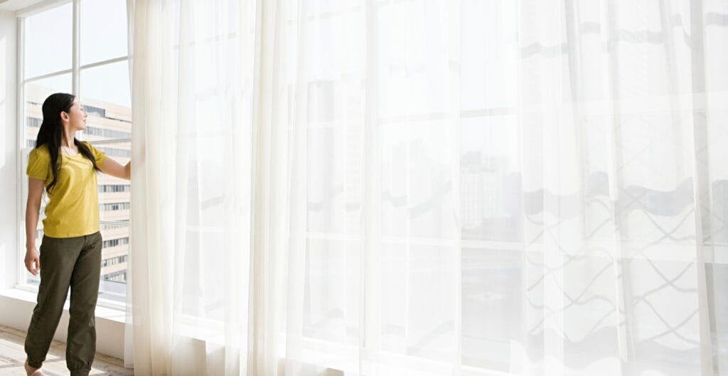 A woman in a yellow top and brown pants is standing by a large window with sheer white curtains, looking outside at a cityscape.