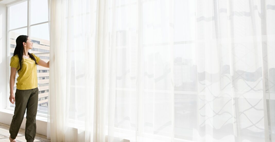 A woman in a yellow top and brown pants is standing by a large window with sheer white curtains, looking outside at a cityscape.