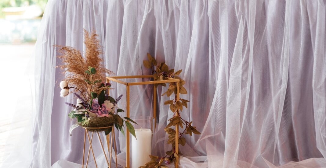 A floral arrangement with dried and fresh flowers displayed in a gold geometric vase, placed next to a white candle, against a backdrop of soft, sheer pink fabric.