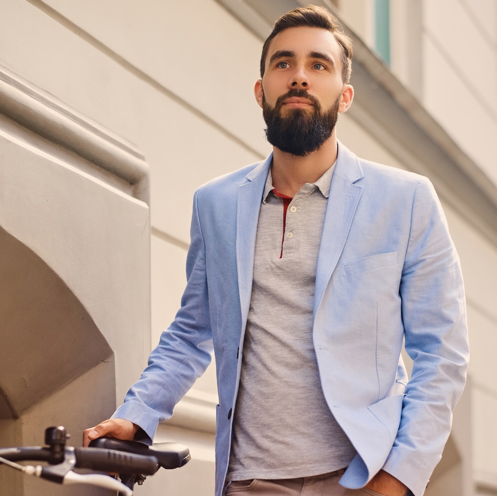 A man wearing a light blue sport coat over a grey polo shirt, standing outdoors with his hand on a bicycle handlebar.
