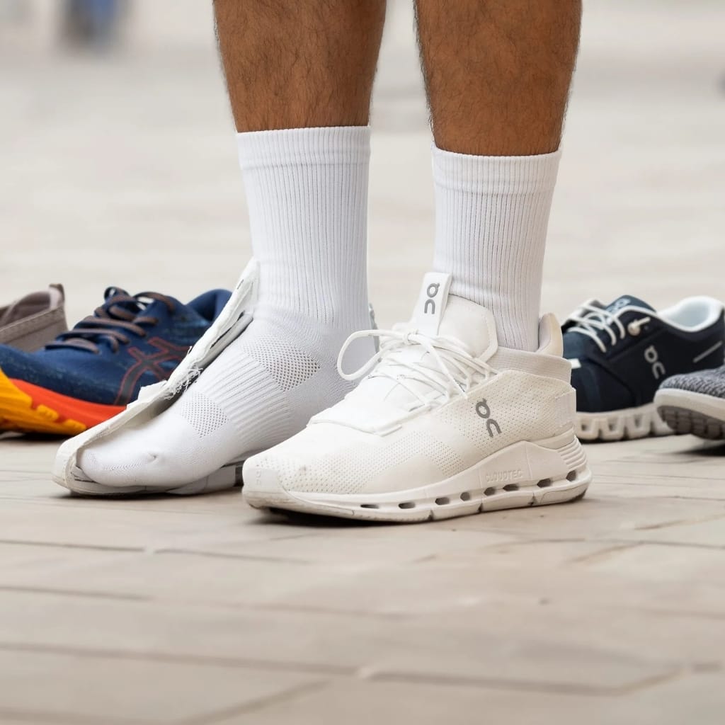 A close-up of a person's legs, showing white athletic socks and white running shoes standing on a light wooden floor. Several other pairs of sports shoes in different colors are visible in the background.
