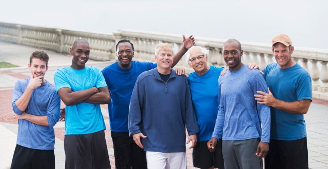 A group of seven men dressed in various shades of blue athletic wear, smiling and standing together outdoors near a railing along a waterfront.