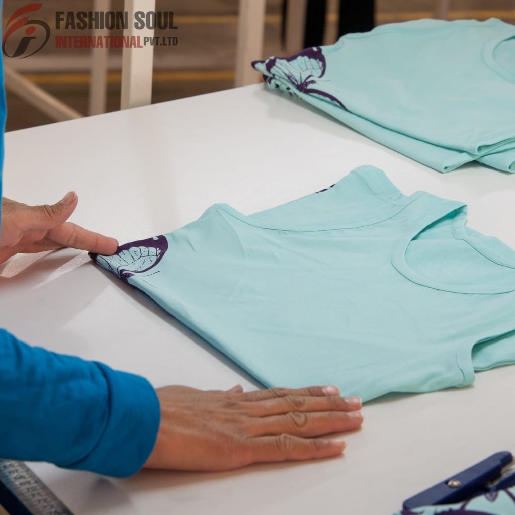 A person measuring light blue fabric with printed designs on a white table, with additional folded fabric in the background.