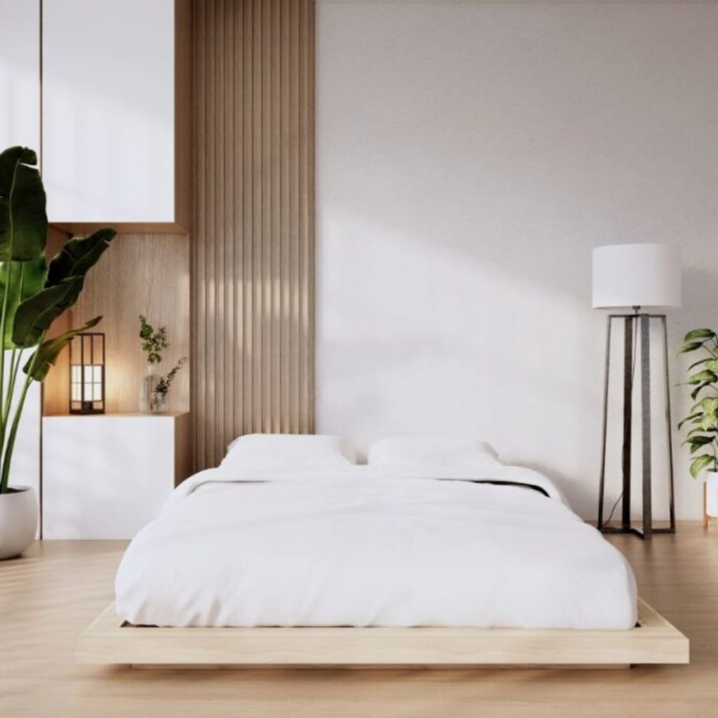 A minimalist bedroom with a low, white bed on a light wooden platform, decorated with white bedding and pillows. The room features a tall black floor lamp, a white nightstand with a small potted plant, and large green plants adding natural elements. Wooden paneling runs vertically behind the bed, creating a warm accent wall.
