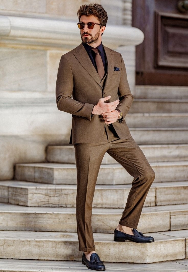 A man wearing a stylish brown suit with a matching sport coat, black shirt, and tie, standing on stone steps outdoors. He is holding his smartphone and wearing sunglasses.