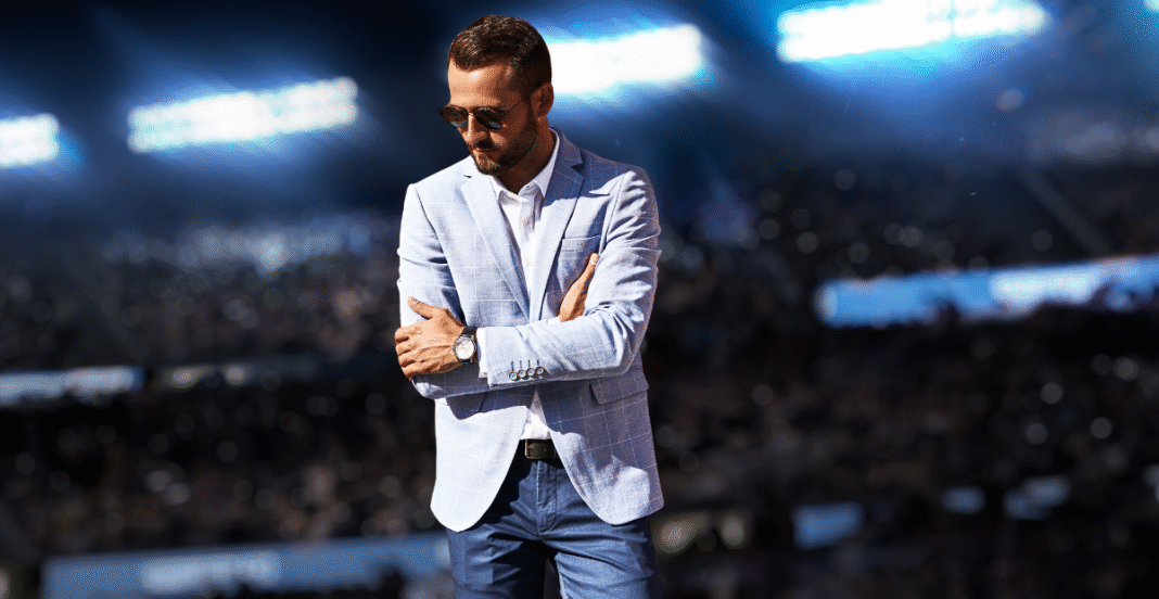 A man wearing a light-colored checked sport coat, white shirt, and jeans, standing with arms crossed in a stadium setting.