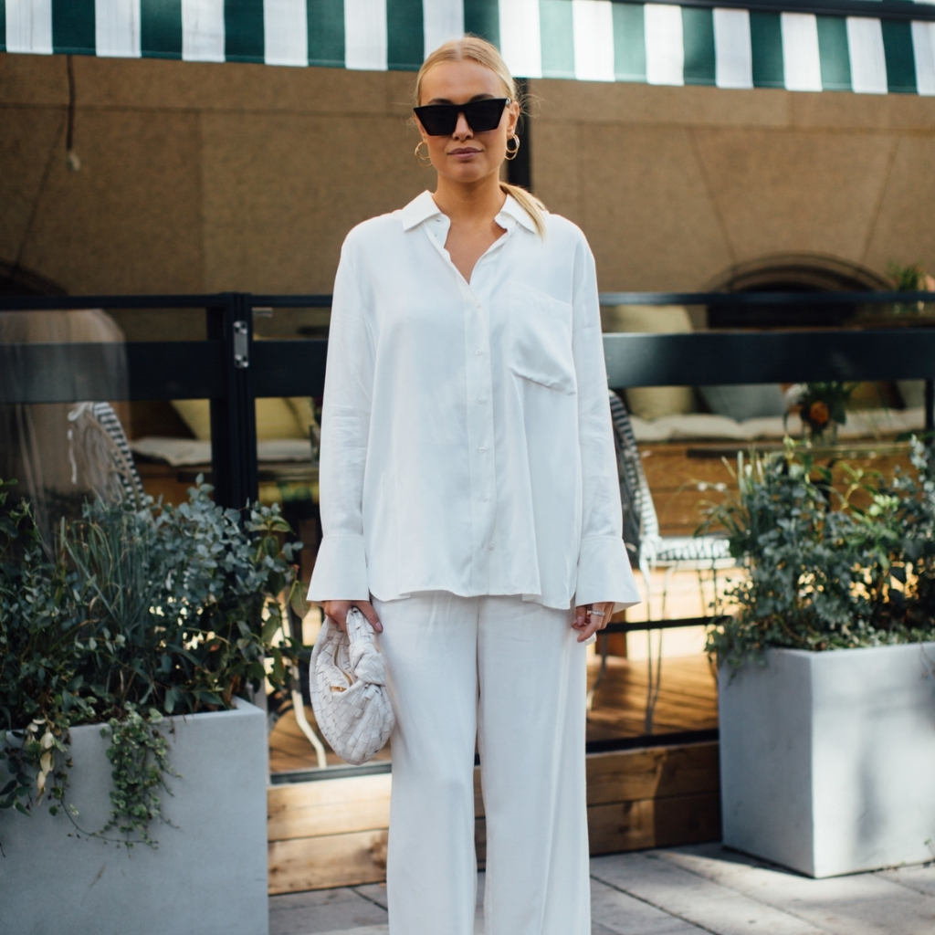 A woman with blonde hair tied back, wearing large black sunglasses, gold hoop earrings, and a loose-fitting white shirt and wide-leg white trousers. She is holding a textured white clutch bag and standing outdoors in front of potted plants and a building with a striped awning.