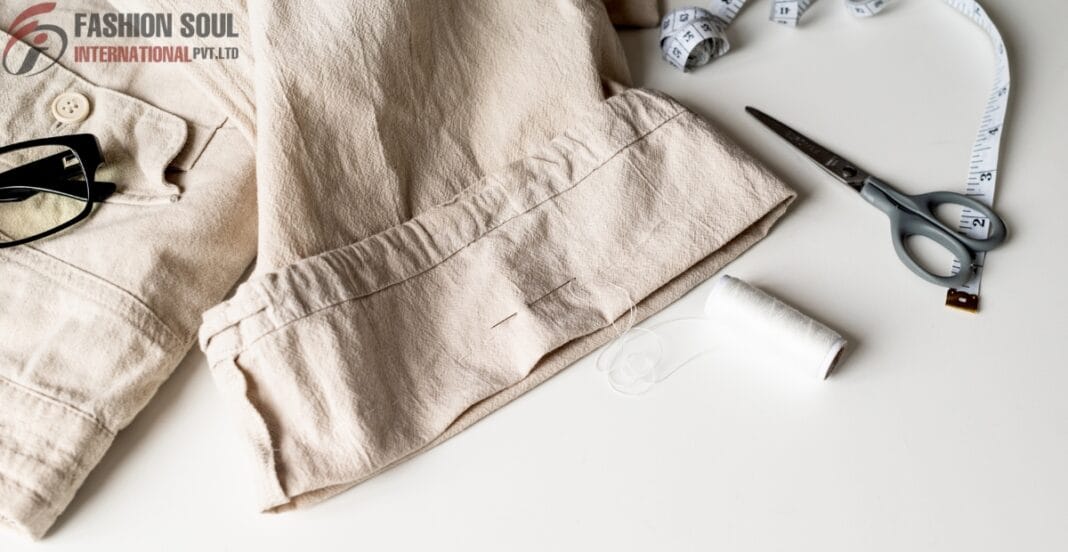 Close-up of beige fabric and sewing materials on a white surface, including a pair of scissors, a measuring tape, a spool of white thread, and part of a beige garment with a pocket, with a 