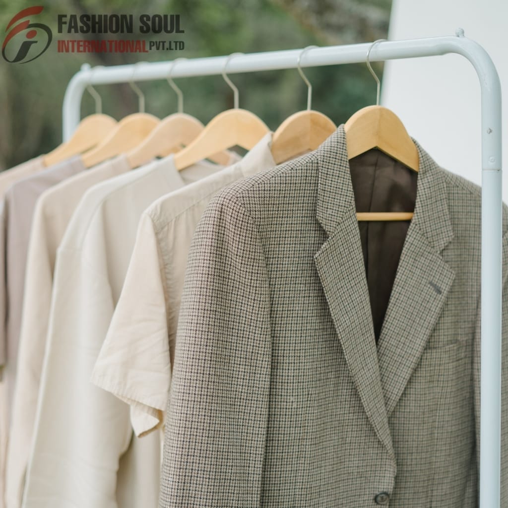 A row of light-colored clothing, including a checked blazer, hanging on wooden hangers on a white rack outdoors, with a blurred natural background.