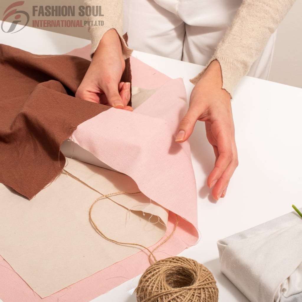 A person is carefully pinning or cutting fabric swatches in shades of brown and pink on a workspace with a white table, with various fabric pieces and a ball of twine nearby.