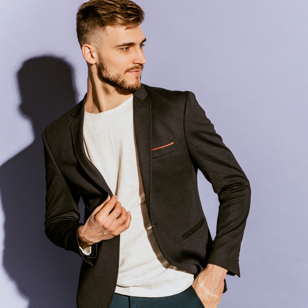 A young man wearing a white crew neck T-shirt paired with a black sport coat, standing against a plain background with his hand holding the lapel of the coat.