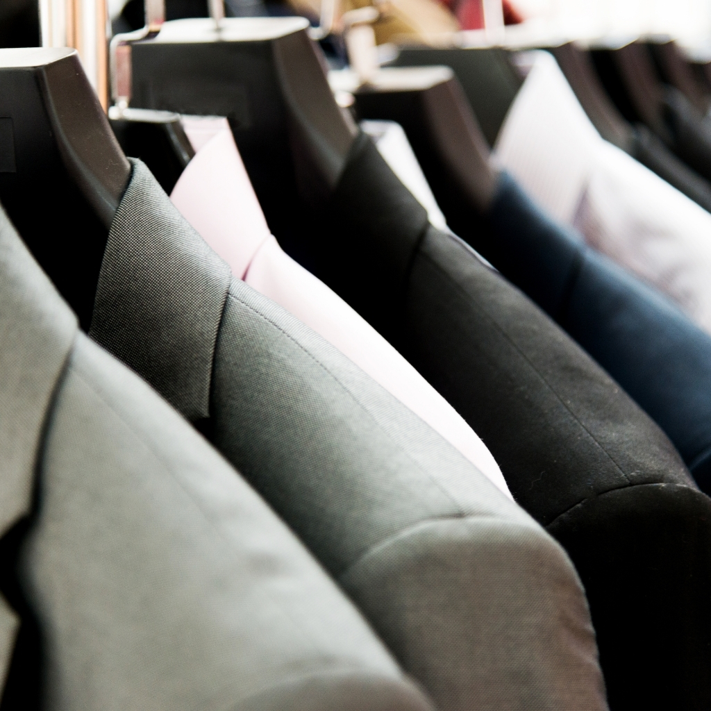 A row of hanging suits on black hangers, in various shades of gray, black, and a hint of pink, displayed closely together in a clothing store.