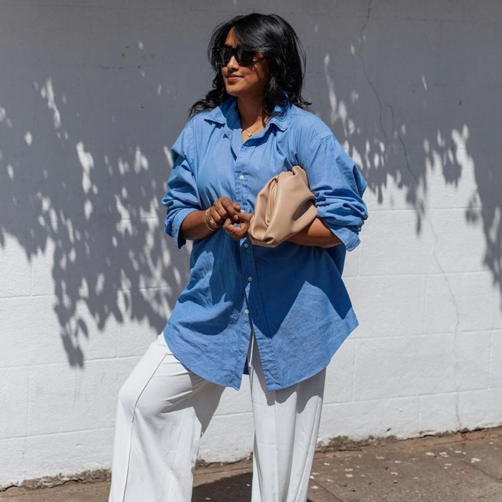  A woman with black wavy hair and sunglasses stands outdoors against a white wall with shadows of leaves, dressed in a loose blue button-up shirt and white wide-leg pants, holding a beige clutch bag.