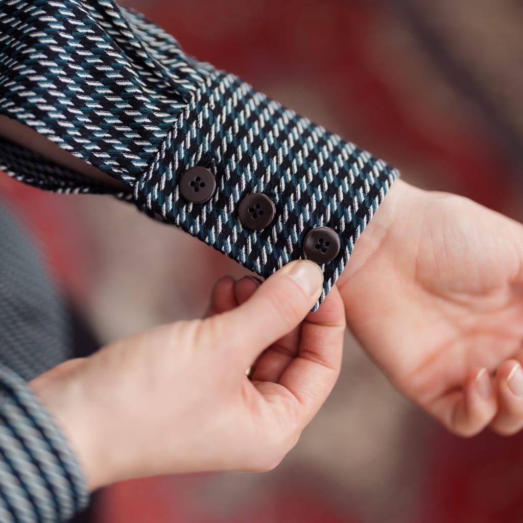 A close-up of a person's hand adjusting the cuff of a shirt with a patterned fabric featuring black, white, and blue stripes, with three black buttons.