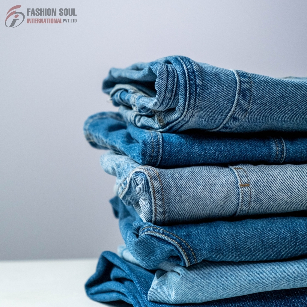 A neatly folded stack of various blue denim jeans placed on a white surface against a plain background. The jeans range in shades from light to dark blue.