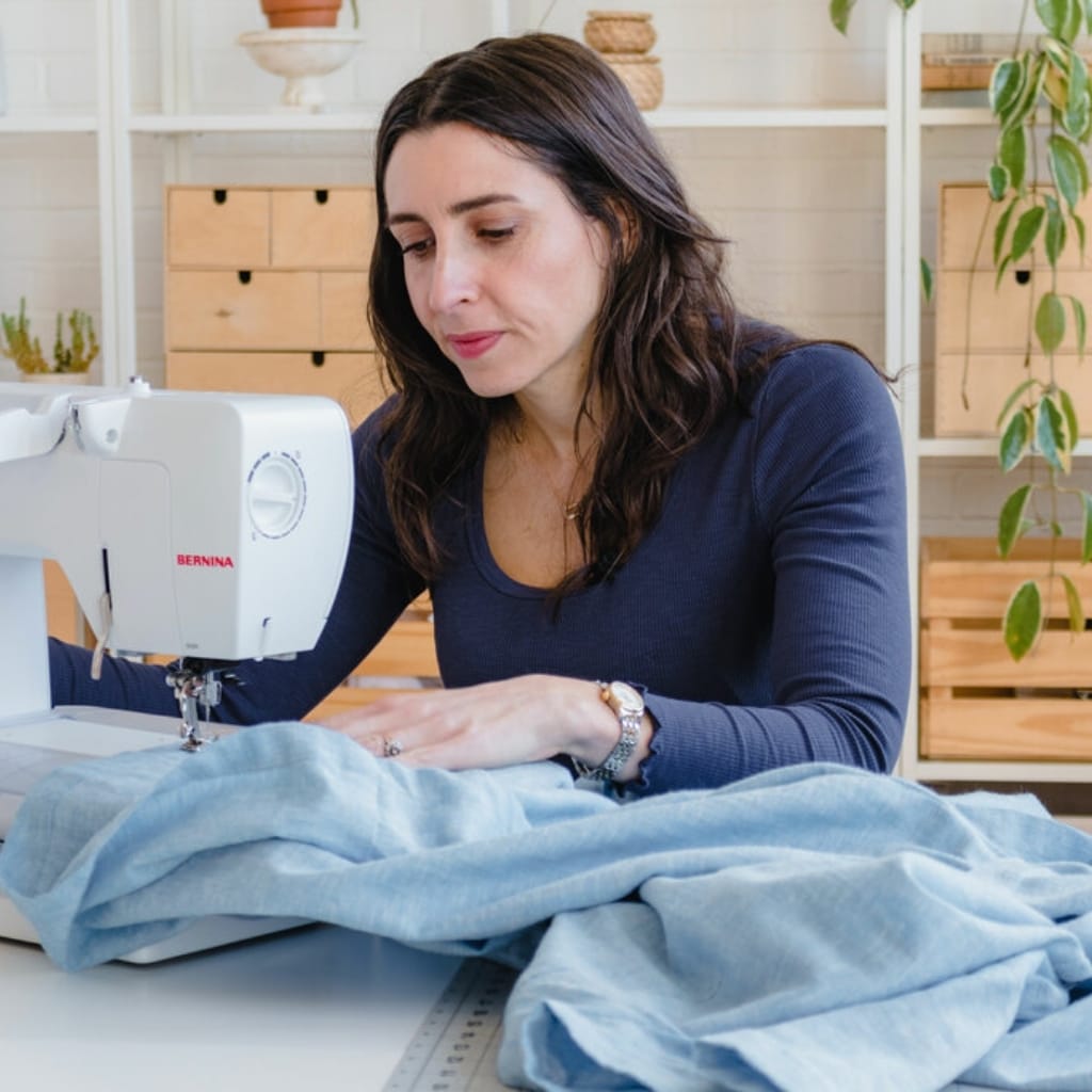 Woman sewing fabric with a sewing machine in a well-lit room with shelves and plants in the background.