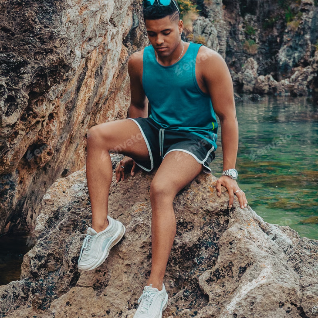 A young man wearing a sleeveless blue athletic top, black shorts with white trim, and white sneakers, sitting on a rocky surface near a body of water in a natural outdoor setting.