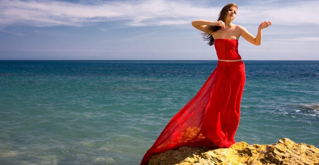 A woman in a red two-piece dress stands on a rock by the ocean, with the sea and sky in the background.