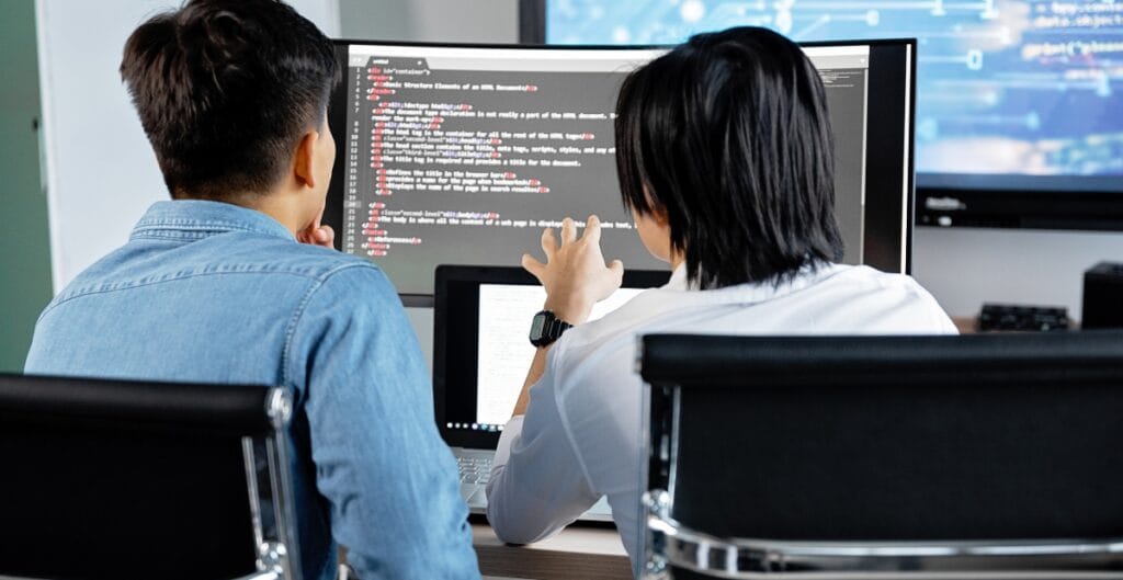 Two people working together in front of multiple computer monitors, one explaining or collaborating on code.