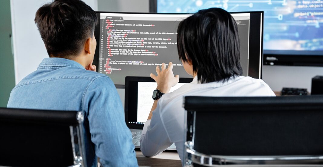 Two people working together in front of multiple computer monitors, one explaining or collaborating on code.