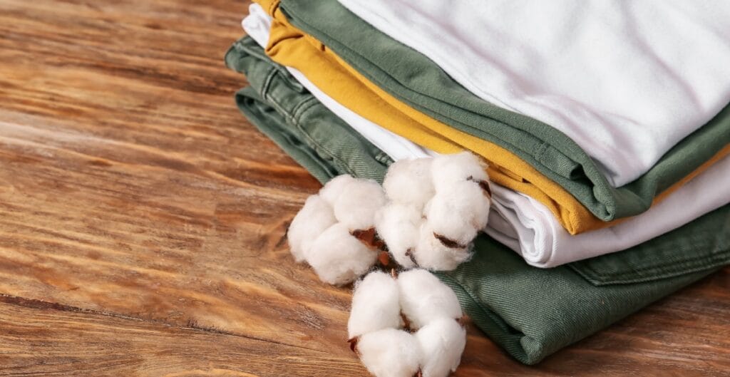 A neatly folded stack of casual clothes, including green, white, and mustard yellow shirts, placed on a wooden surface. Cotton flowers are casually arranged beside the clothes.