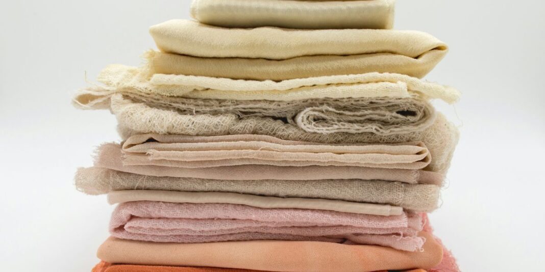 A stack of folded, natural-dyed fabric in soft, earthy tones ranging from cream and beige to blush and rust.