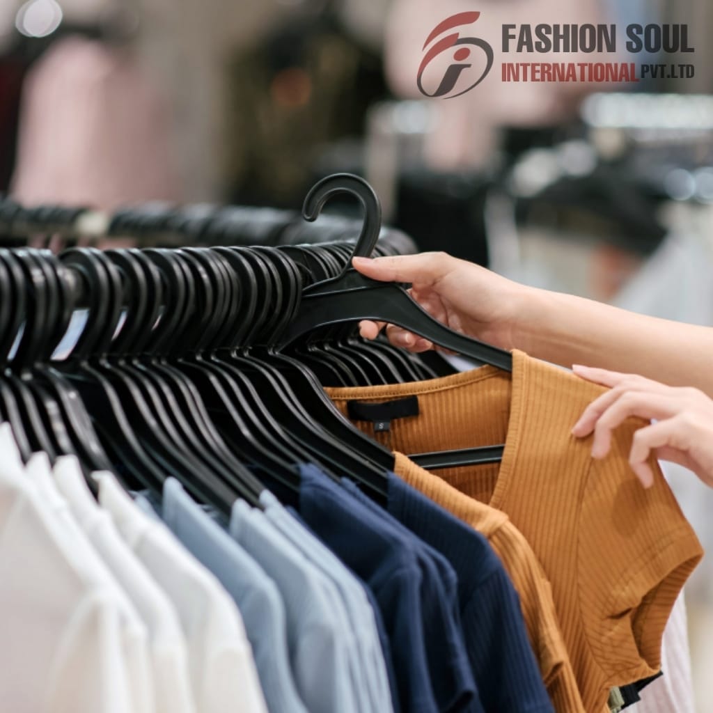 A person shopping for clothes, holding a mustard-colored ribbed top while browsing a rack of various colored shirts.
