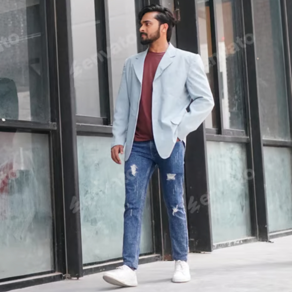 A man walking outdoors, dressed in a light gray sport coat, rusty-red T-shirt, distressed blue jeans, and white sneakers.