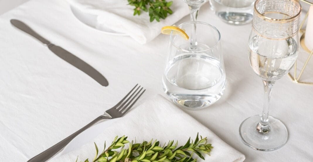 A close-up of a elegantly set table with a white tablecloth, a fork on the left, a knife on the right, a sprig of greenery, a glass of water with a lemon wedge, a water pitcher, a wine glass, and some decorative elements in the background.